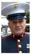 Marine in uniform, wearing a white hat, looks slightly off-camera. Red trim on collar, gold buttons.