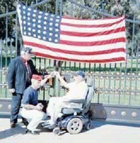 Three men raise glasses in front of an American flag and gate; one sits in a wheelchair.