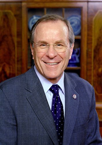Man in suit, smiling, wearing glasses. Indoors, wooden cabinet in background.