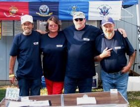 Four people in matching blue shirts pose in front of military flags.