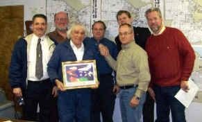 Group of men, indoors, holding a framed picture. One man points, others smile. Maps are in background.