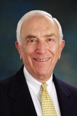 Smiling older man in a suit and tie, against a blurred blue background.