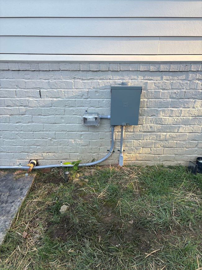 Gray electrical boxes and conduit on a brick house exterior above a patchy grassy yard