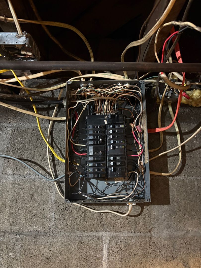 Open electrical breaker panel with tangled wires in a cinder block basement wall