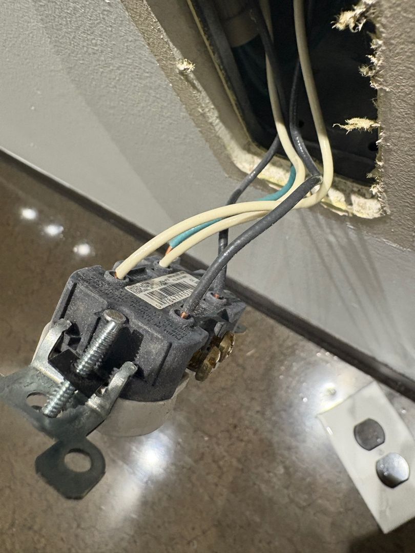 Exposed electrical outlet wiring protruding from a wall opening, with a loose black outlet hanging nearby