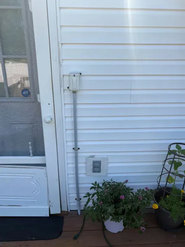 White house exterior with a door, window, utility conduit, and potted plants beside a black mat.