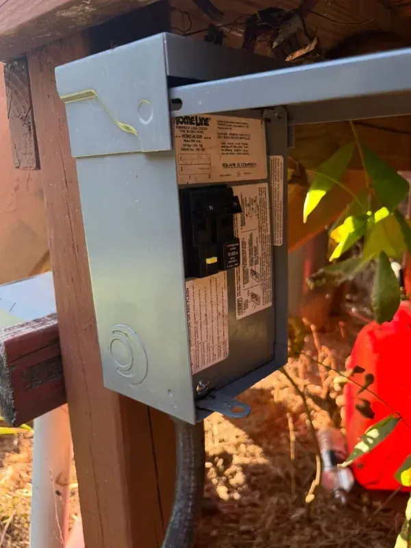Outdoor electrical control box mounted on a wooden post beside a vine-covered wall and red flowers