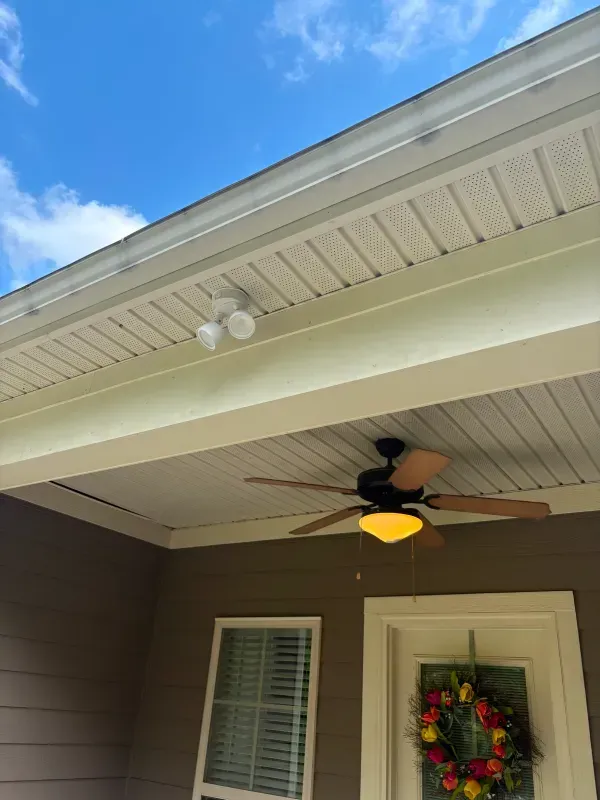 Covered porch ceiling with lit ceiling fan, white trim, and a wreath on the door under a blue sky
