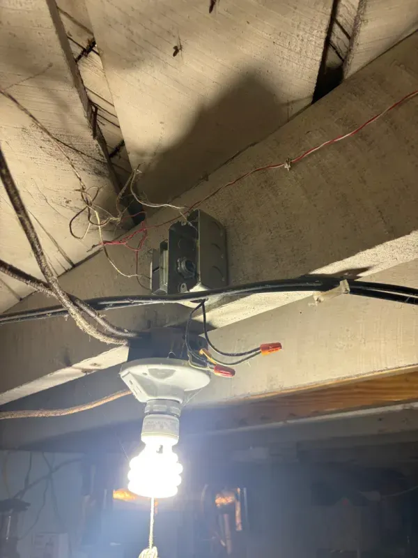 Ceiling fan with a lit bulb hanging from a rough, unfinished ceiling with visible wiring.