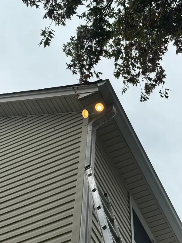 House corner with two lit security lights under the roof eave, viewed against a cloudy sky and tree branches