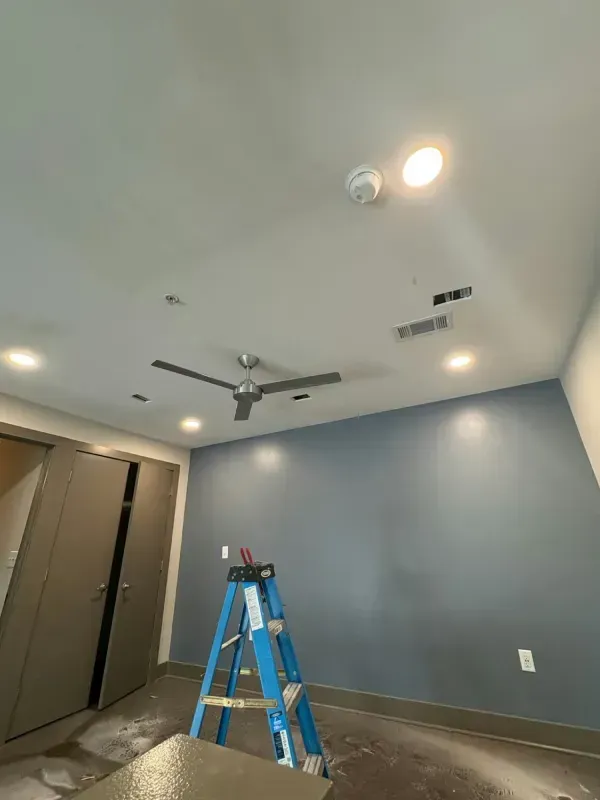 Empty room with blue-gray walls, ceiling fan, lights, and a blue step ladder on the floor