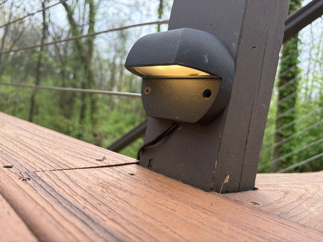Small wooden doll with a black cap peeks from a railing on a deck outdoors.