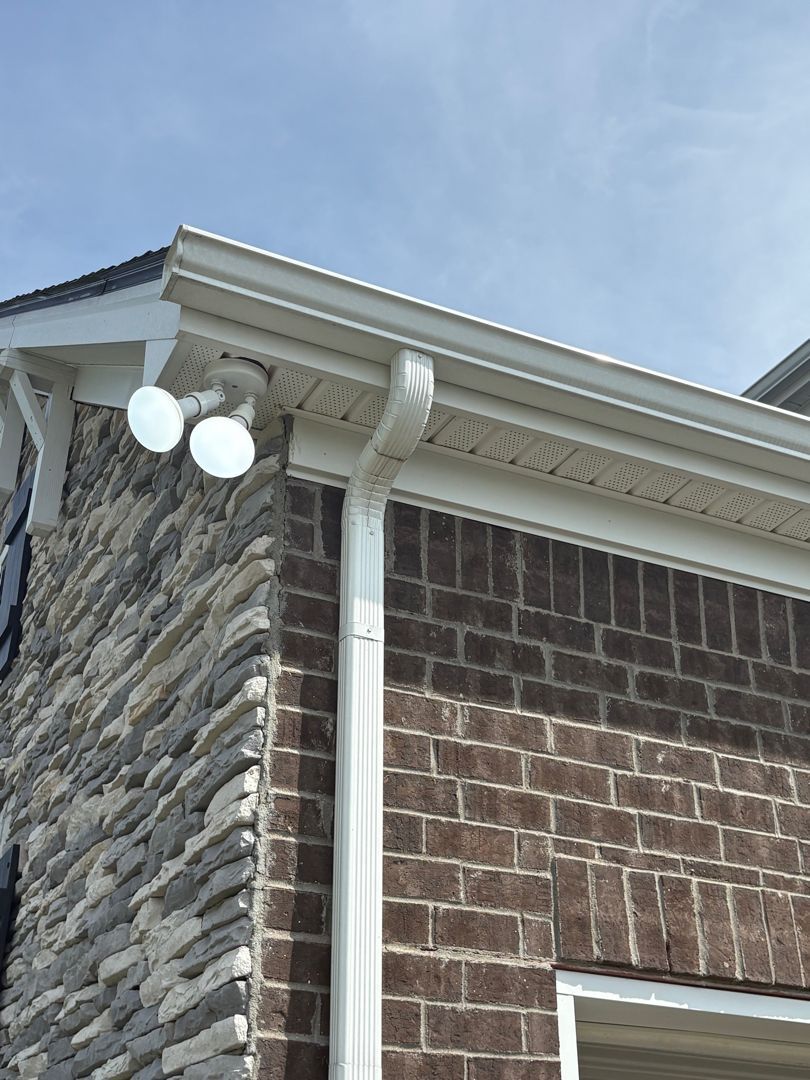 Stone-and-brick house corner with white gutter and downspout under a blue sky.