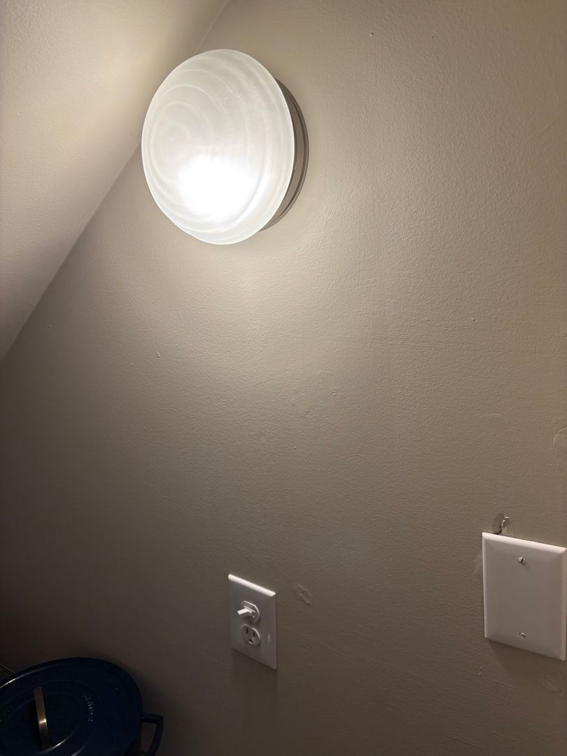Wall with round ceiling light, outlet, and switch plate on beige paint.