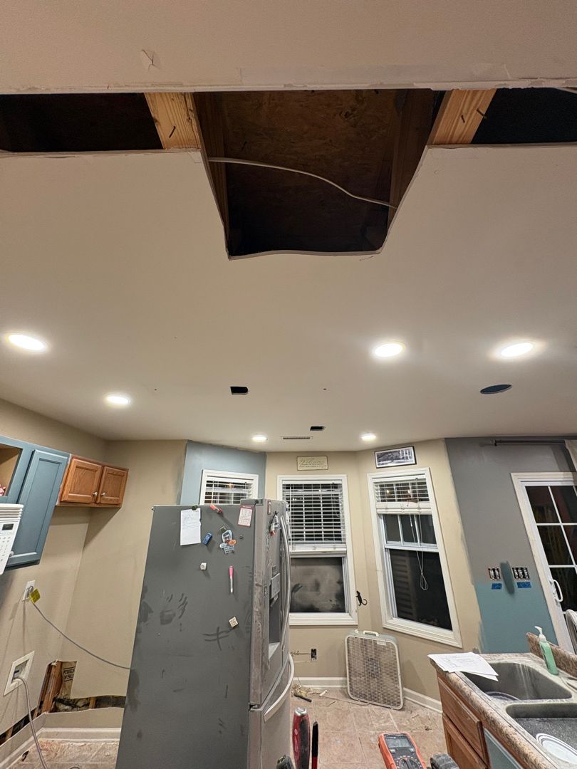 Kitchen with damaged ceiling opening above a gray refrigerator and cabinets, showing a partially exposed attic space.