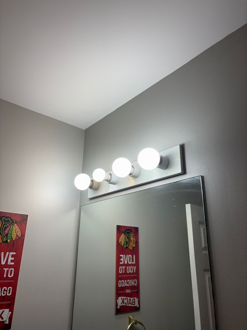 Bathroom mirror with four lights above it and a gray wall corner reflected in the mirror.