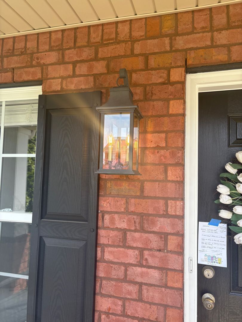 Brick house entryway with black door, wall lantern, and white-framed window, decorated with a wreath