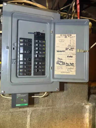 Open electrical breaker panel with labeled circuits and gray metal door on a basement wall