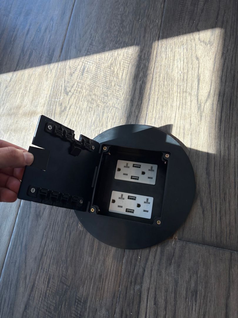 Hand opening a black wall-mounted outlet cover beside two electrical outlets on a wooden floor