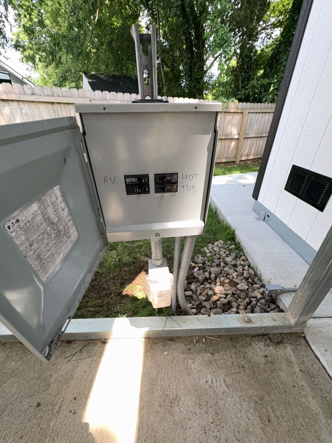Outdoor electrical service panel beside a house, with open gray cover and gravel base.