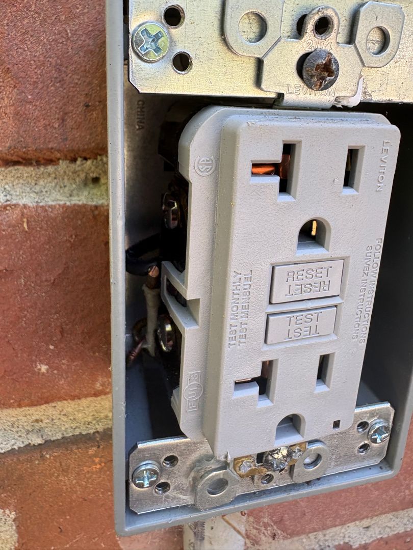 GFCI electrical outlet mounted in a metal box on a brick wall, with wiring exposed.