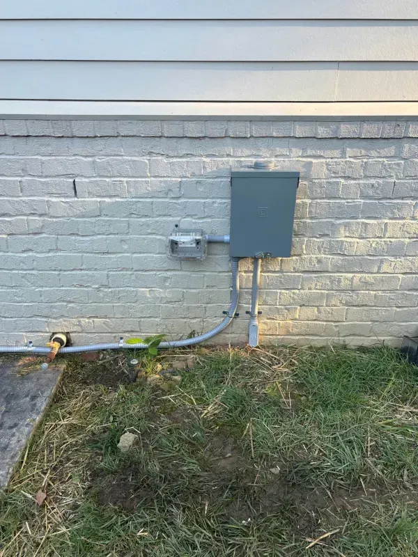 Gray utility boxes and conduit mounted on a white brick exterior wall above patchy grass.