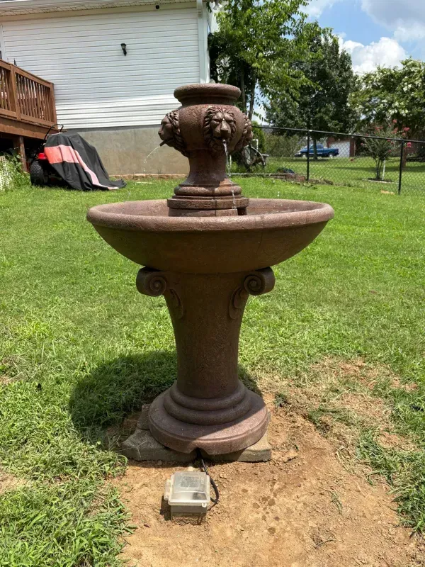 Rust-colored outdoor fountain on a concrete base in a grassy yard near a house.