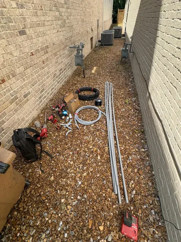 Work tools and pipes laid out on gravel between two brick walls in a narrow side yard