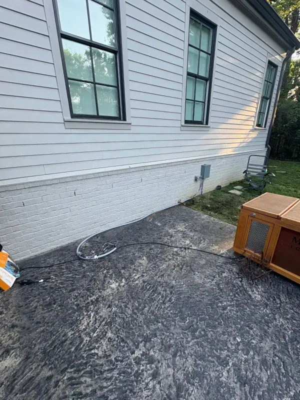 Side of a white house with windows, concrete foundation, and a dark paved patio with a small wooden crate.