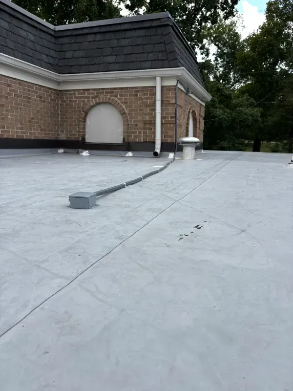 Flat rooftop with a white membrane, brick wall, and a low parapet near a tree-lined background.