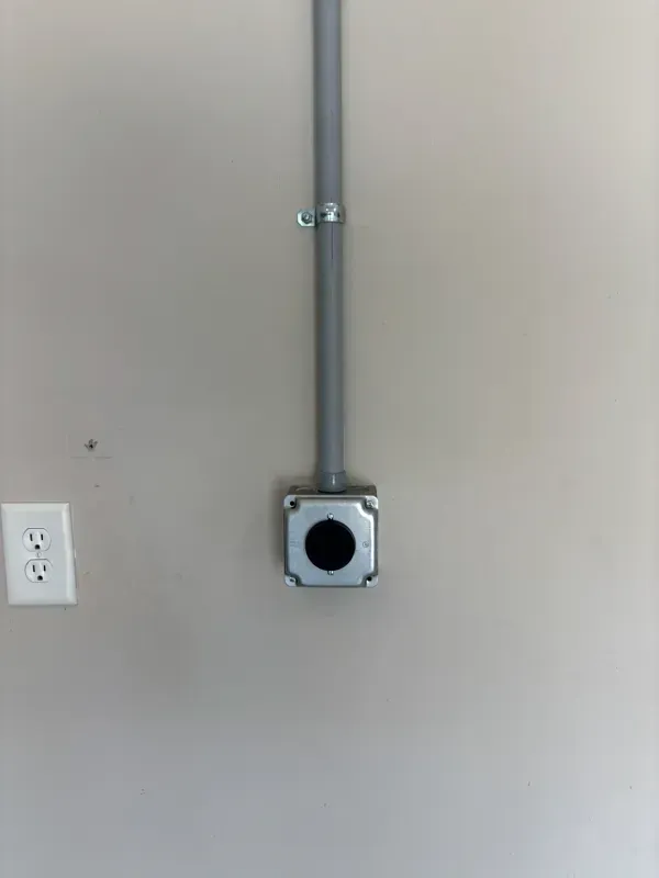 Gray electrical conduit leading to a metal junction box on a beige wall, with an outlet on the left.