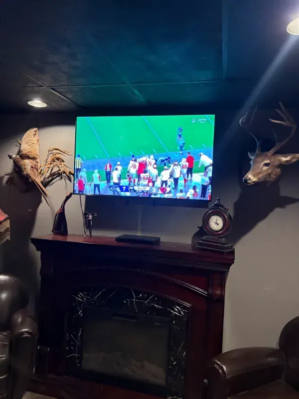 Living room with a TV showing a soccer match above a fireplace, flanked by deer heads and lamps.