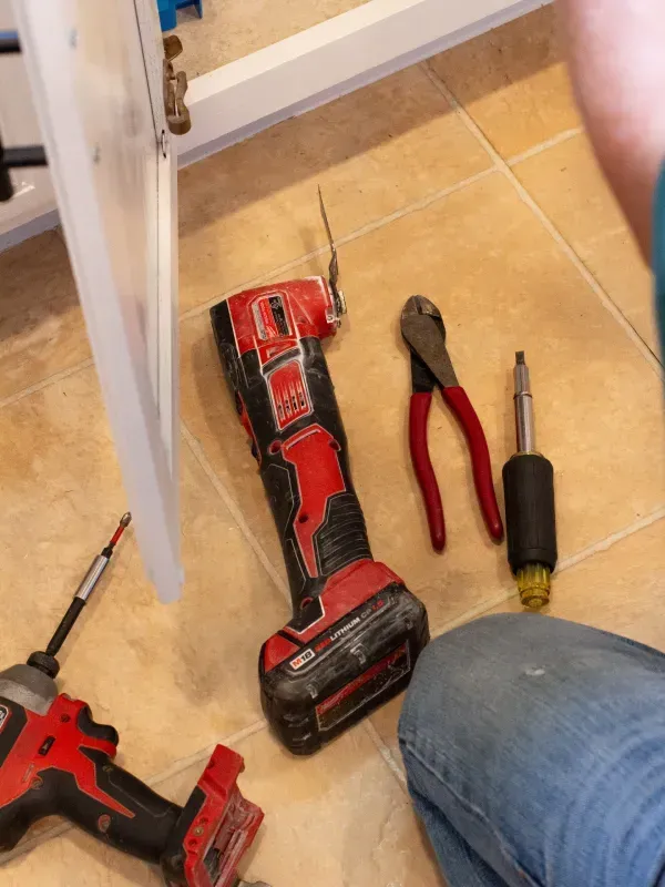 Red cordless drill, pliers, screwdriver, and another tool on beige tile floor during home repair