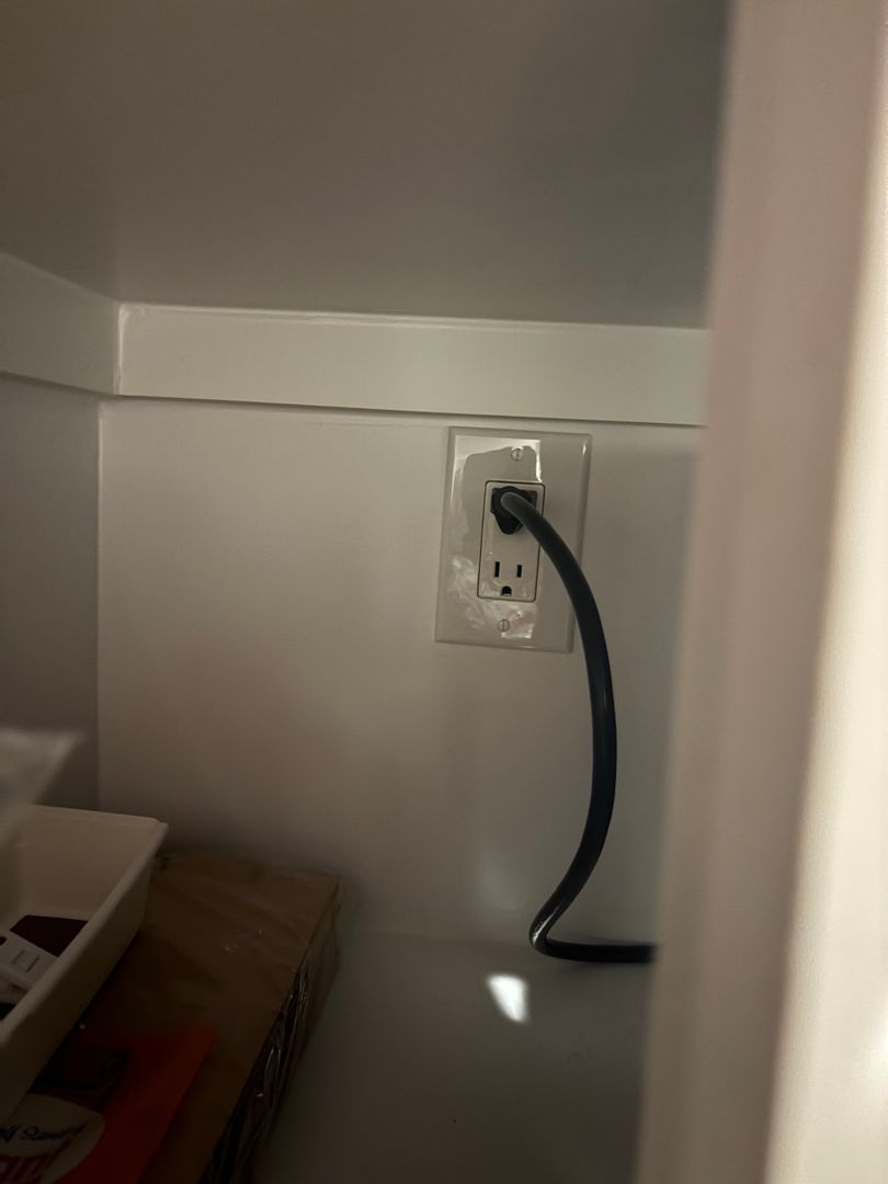 White wall outlet with a black cord plugged in beside a closet or cabinet opening.