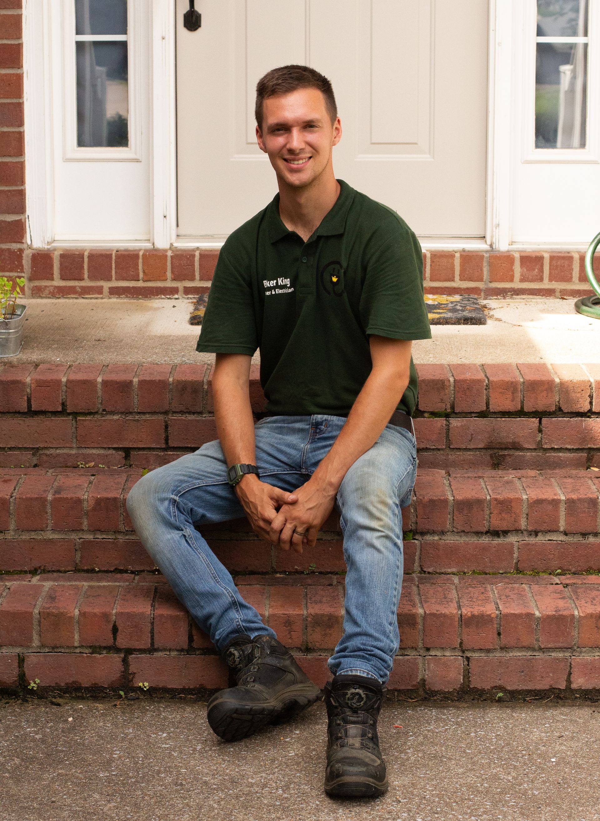Young man sitting on brick steps outside a white door, wearing a dark green shirt and jeans, smiling.
