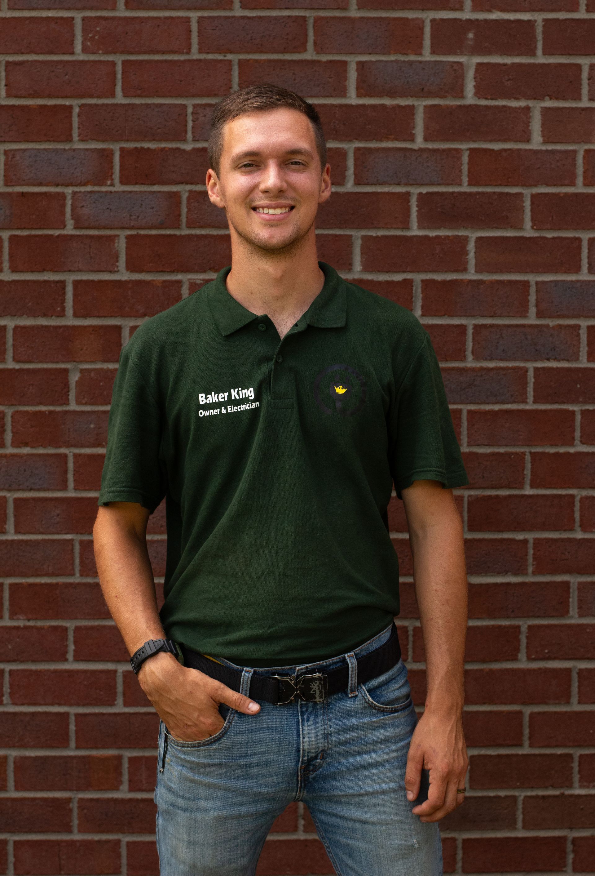 Standing against a brick wall, a smiling person wears a dark green polo and jeans.