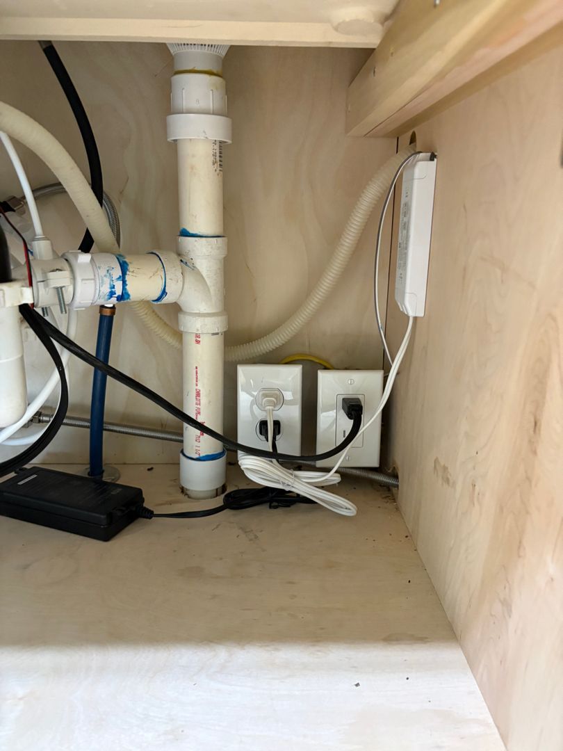 Under-sink cabinet with white PVC plumbing, cables, and an outlet panel inside a wooden cabinet