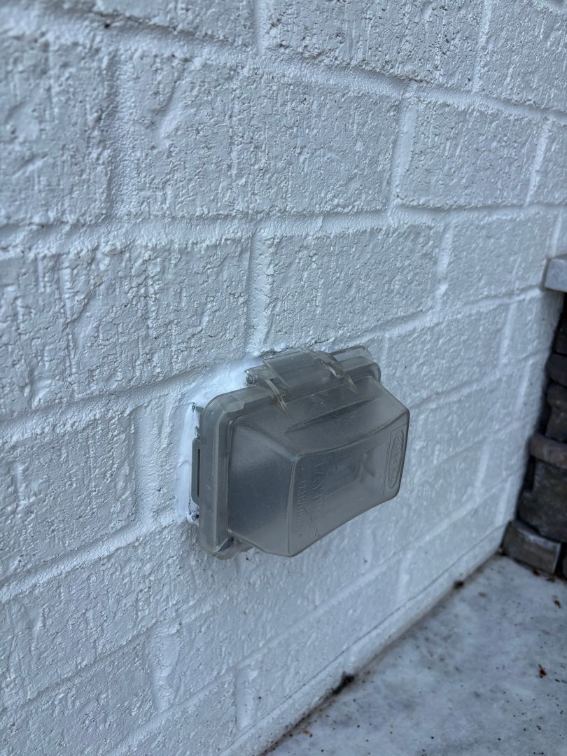 Gray outdoor electrical box mounted on a white brick wall, partly covered with tape or sealant.