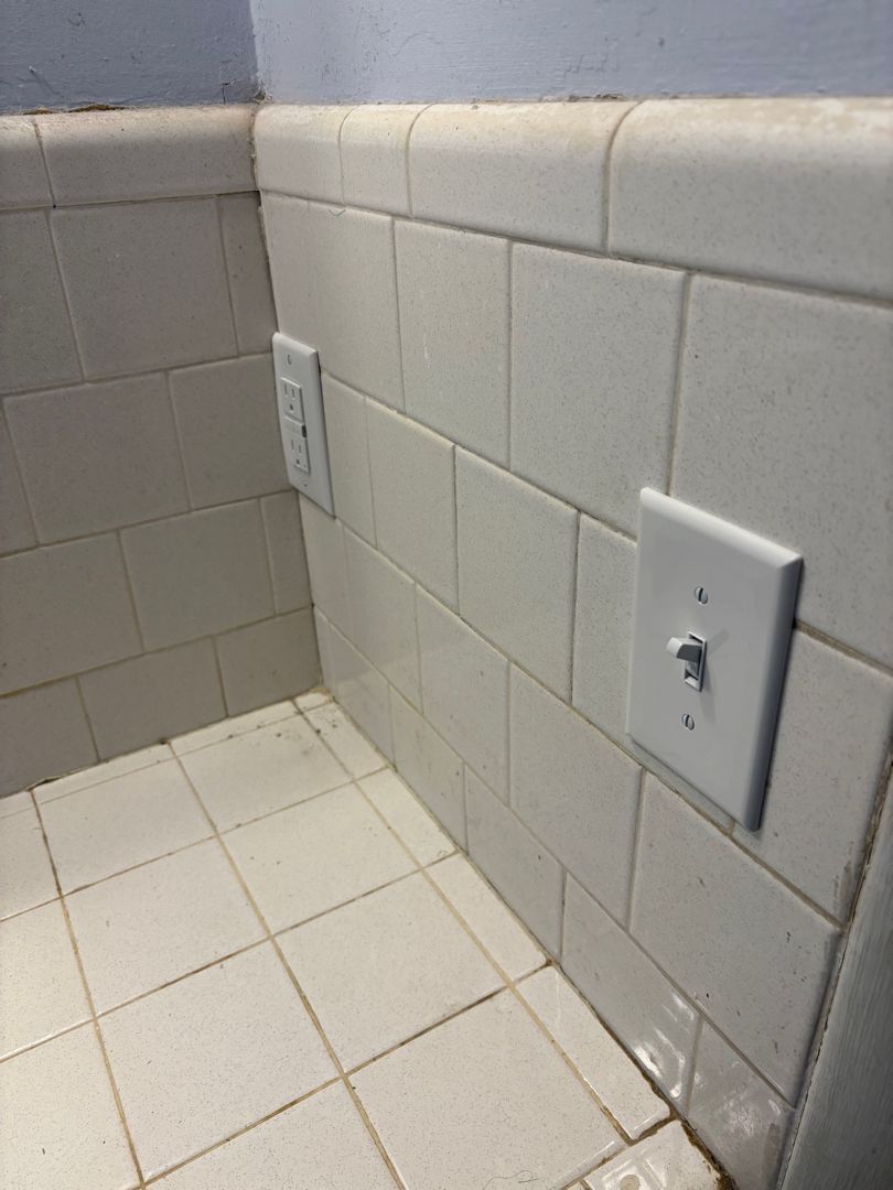White tiled bathroom corner with two wall outlets and a baseboard seam near the floor
