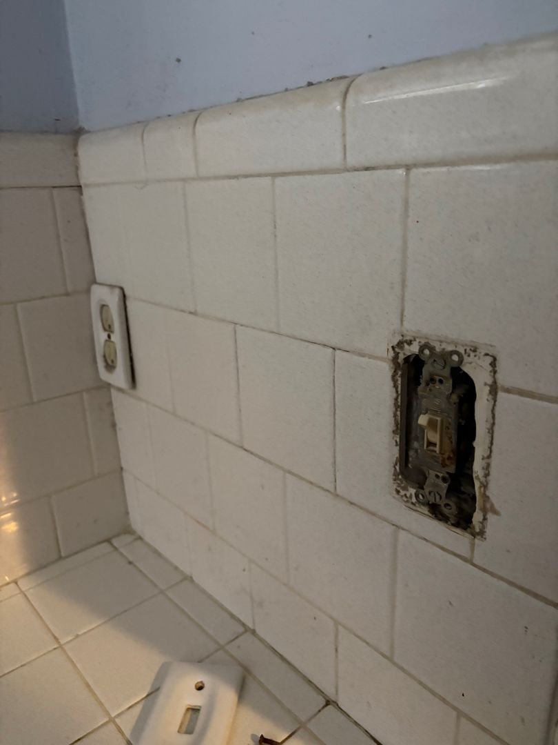 Dirty white tiled wall with two exposed electrical outlets and a sink corner below.