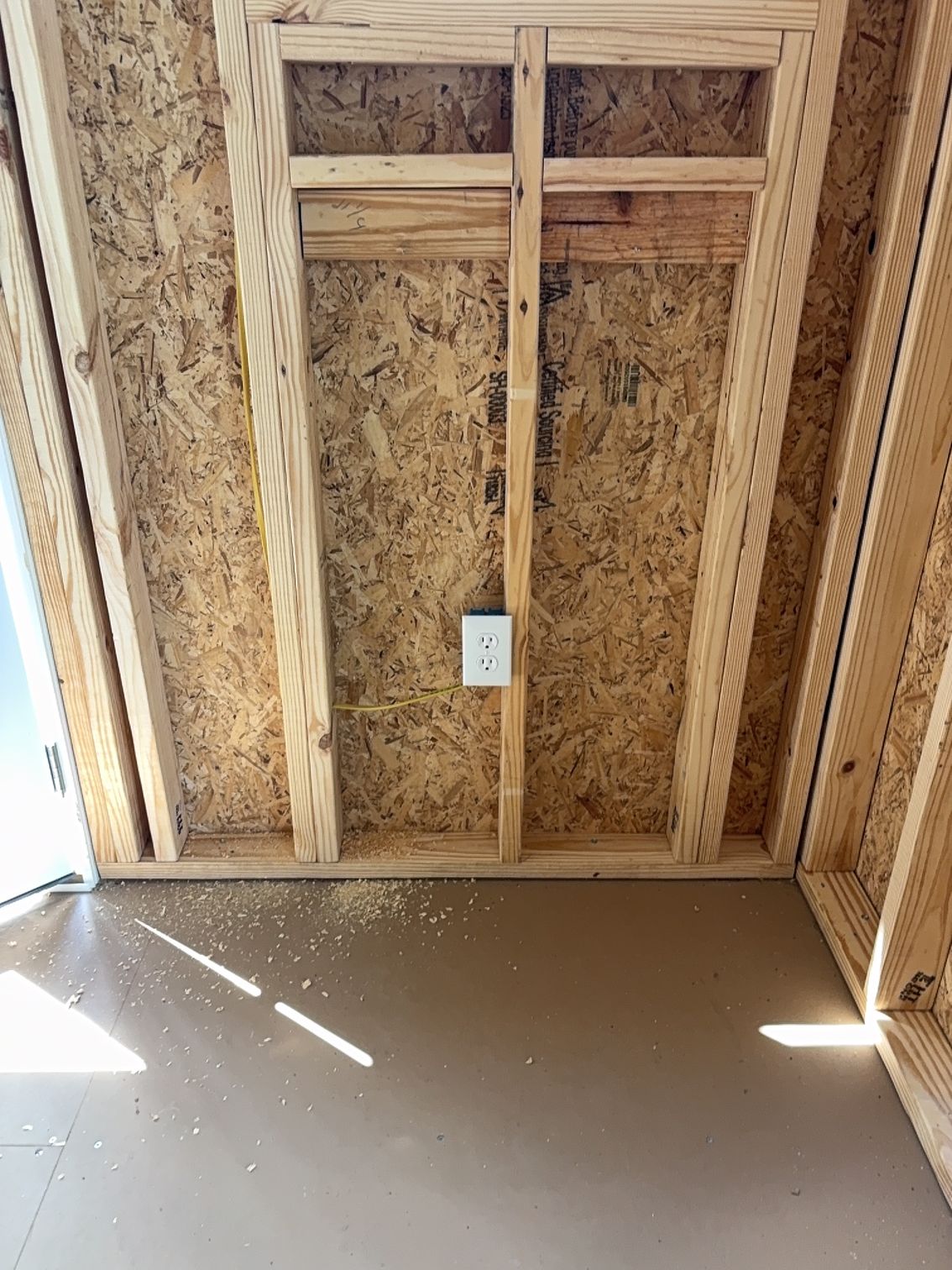 Unfinished wall framing with exposed wood studs and a single electrical outlet, above a brown subfloor.
