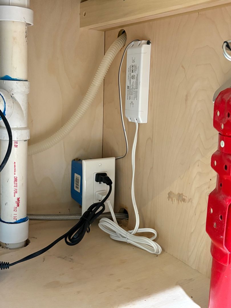 Under-sink cabinet with white pipes, electrical outlets, and coiled cords beside a red storage bin.