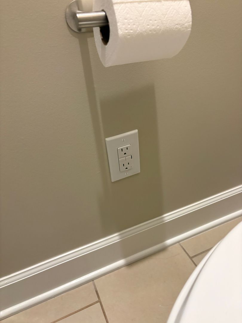 Bathroom wall with toilet paper holder, electrical outlet, and white baseboard tile floor