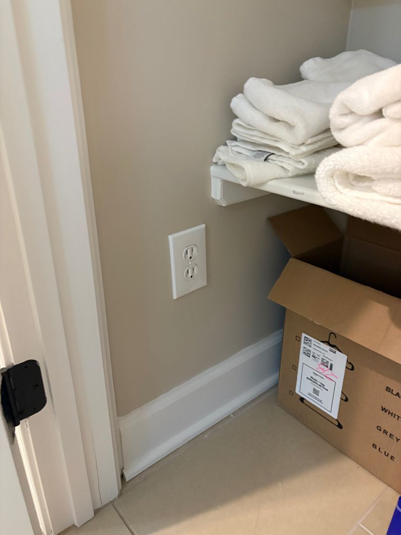 Closet corner with folded white towels on a shelf, a cardboard box, and an electrical outlet near the floor