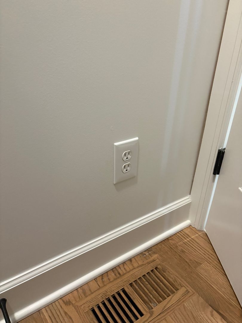 Wall outlet above a floor vent in a beige hallway corner with white trim and hardwood floor.