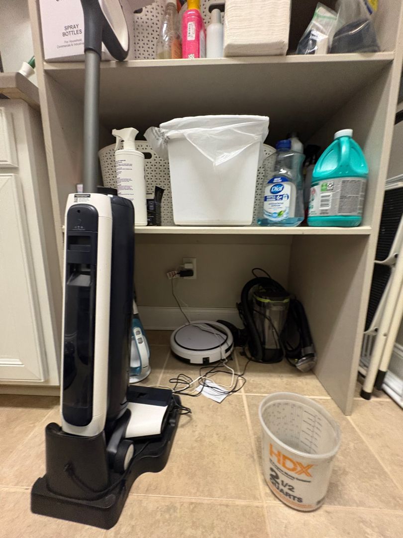 White shelving unit with cleaning supplies, a floor vacuum, and a trash bin on a tiled floor.
