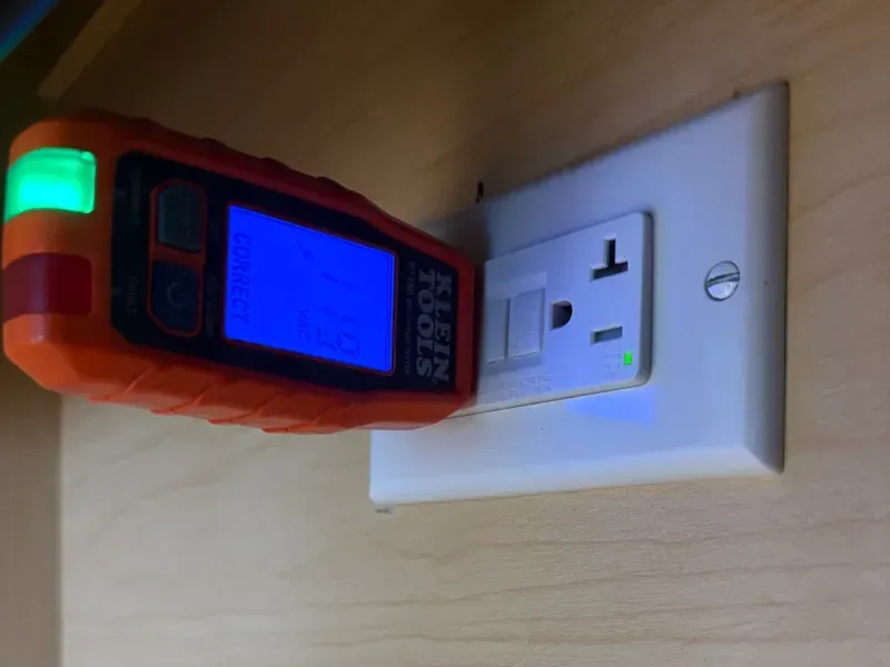 Orange handheld tester plugged into a white wall outlet on a wood surface
