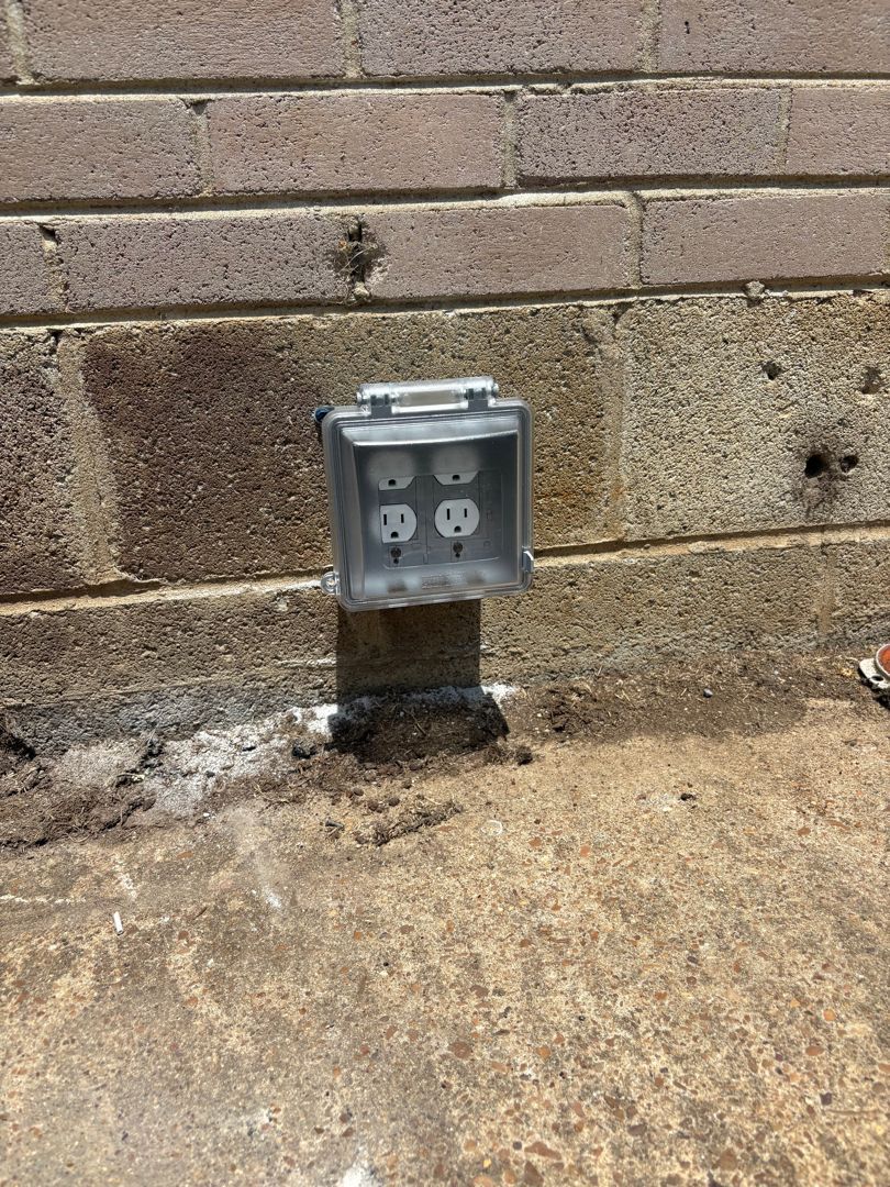 Outdoor electrical outlet in a gray weatherproof box on a brick wall near concrete ground