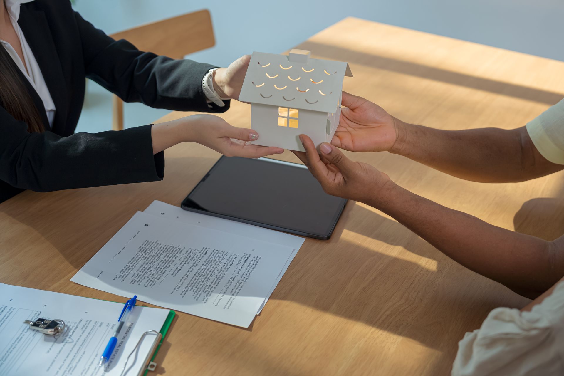 Real estate agent handing a model house to a person at a table, documents and keys present.