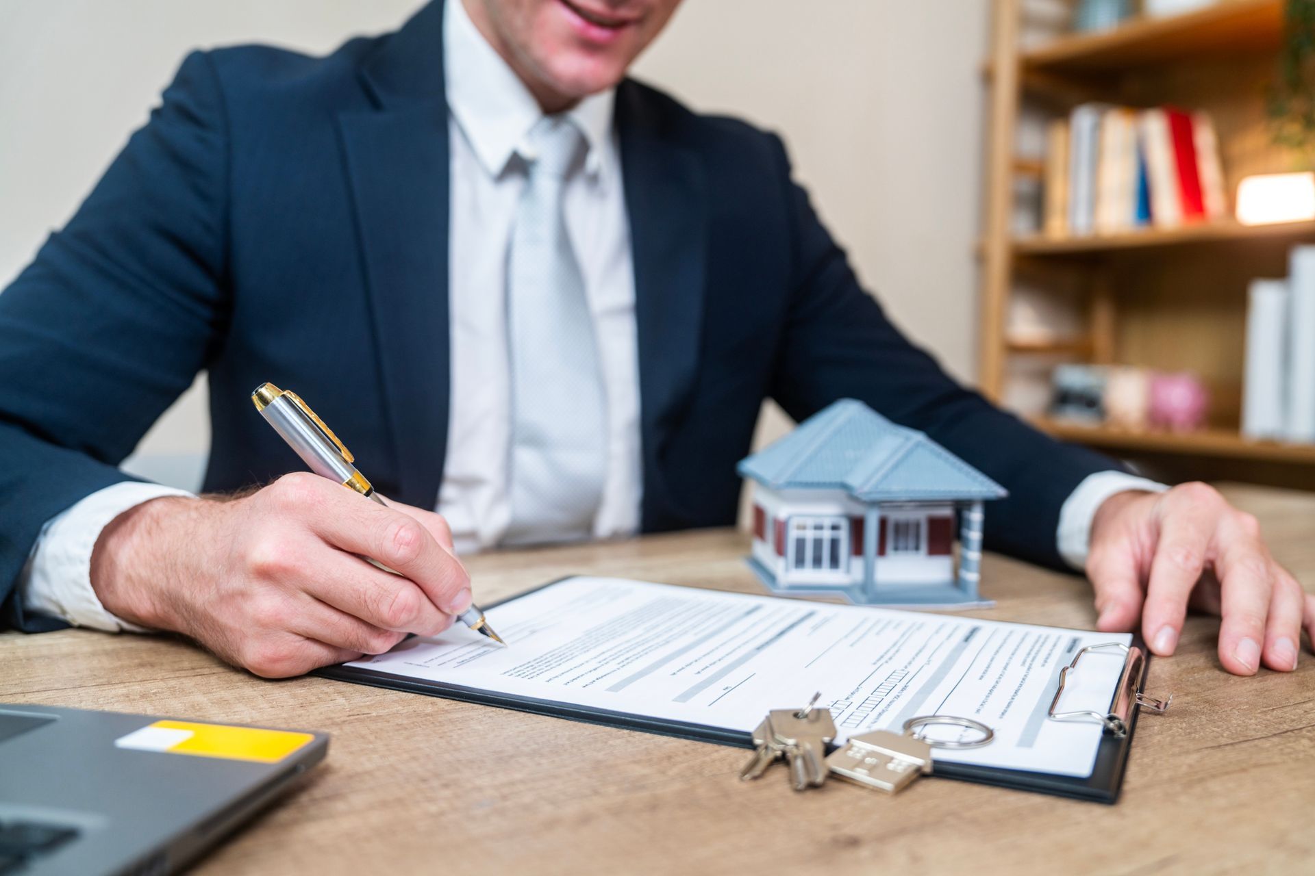 Real estate agent signing documents, small house model and keys on desk.
